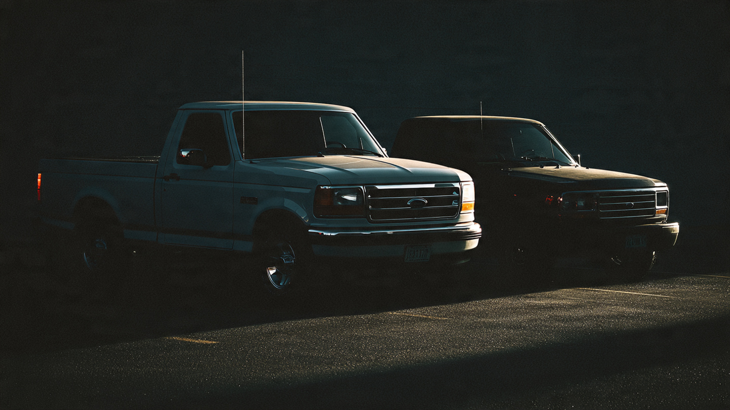 Old compact pickup trucks in a dusty junkyard contrasted against gleaming modern full-size trucks