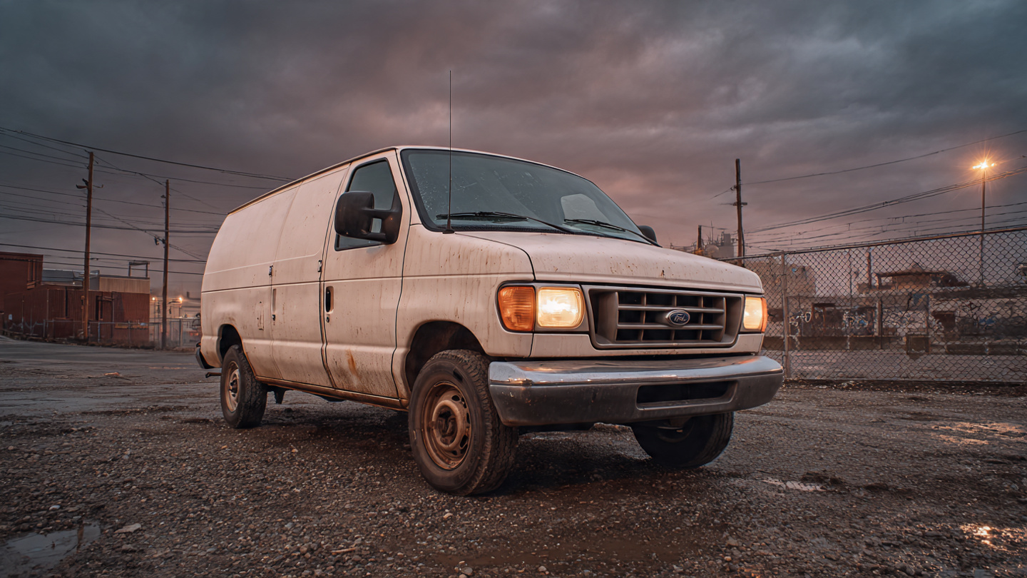 A weathered Ford E-350 commercial van on a gritty industrial lot at dusk