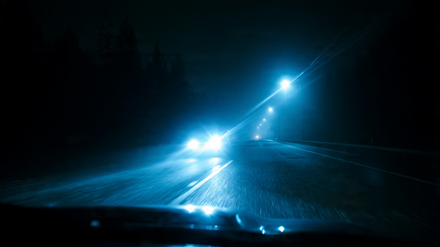 Blinding LED headlights from an oncoming vehicle creating intense glare on a dark two-lane road