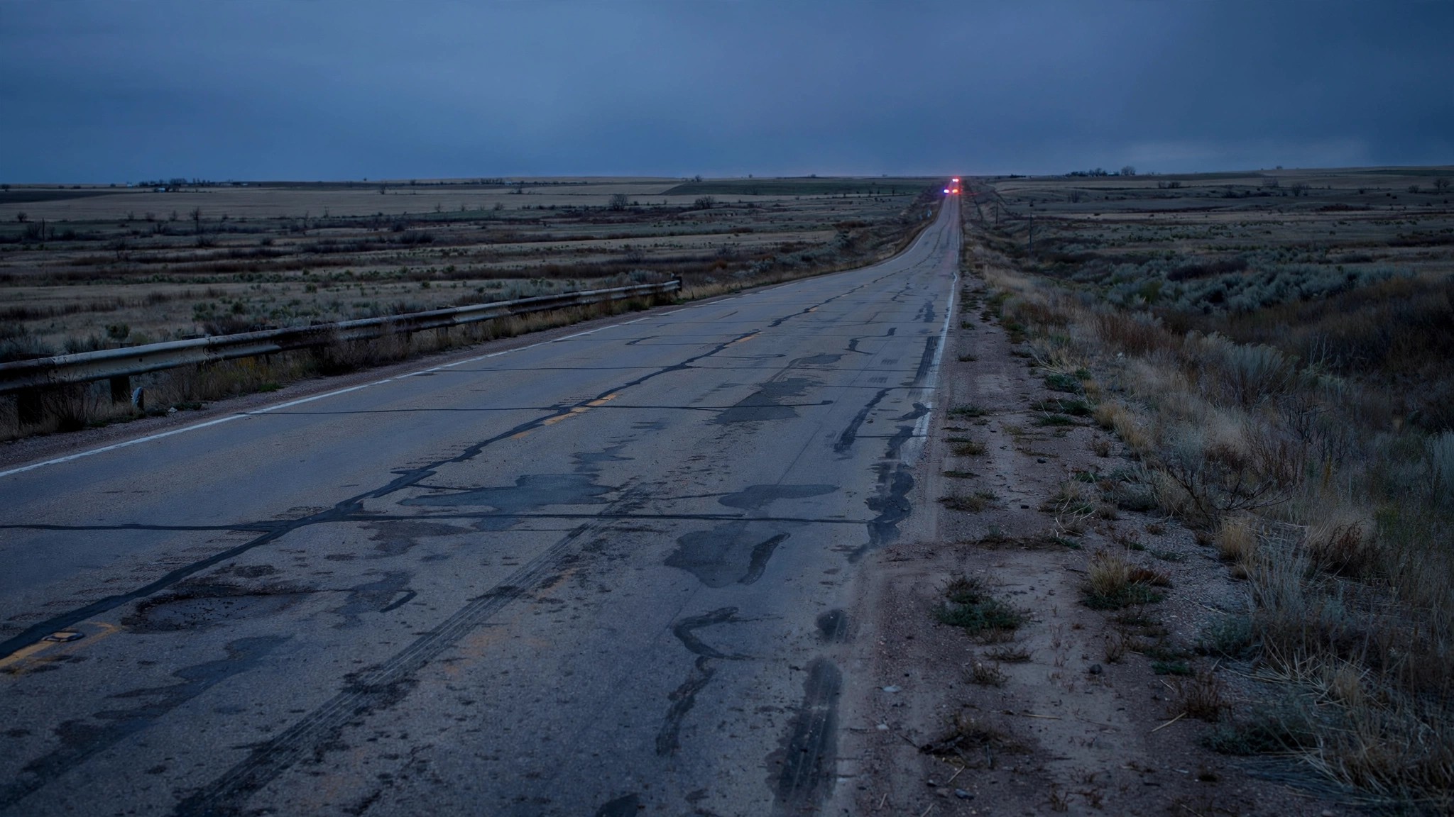 A desolate rural interstate highway stretching into the horizon at dusk, with emergency lights barely visible in the distance